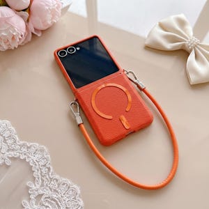 以下が含まれることがあります： 鮮やかなオレンジ色の折りたたみ式スマートフォンケース。表面はテクスチャー加工が施され、背面には円形のマグネット式充電リングが付いています。ケースには同色のオレンジ色のリストストラップが銀色の金属製クリップで取り付けられています。