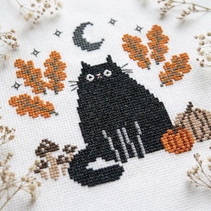 Pode incluir: Um bordado em ponto cruz sobre tecido branco apresentando um gato preto grande sentado centralmente. O design inclui folhas de carvalho laranja, pequenos cogumelos castanhos, duas abóboras e uma lua crescente escura com estrelas.