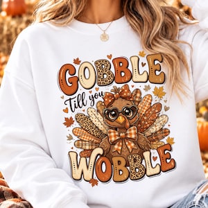 Peut inclure: Un sweat-shirt blanc à manches longues orné d'un motif de dinde portant des lunettes en forme de cœur et un nœud papillon à carreaux. Le texte GOBBLE TILL YOU WOBBLE est imprimé en tons marron et beige, entouré de feuilles d'automne.