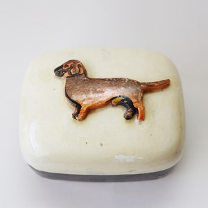 May include: A white ceramic trinket box with a black base. The lid features a brown and black dachshund dog figurine.