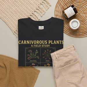Puede incluir: Una camiseta negra con el texto "CARNIVOROUS PLANTS A FIELD STUDY" en letras doradas. Debajo del texto, una ilustración gráfica muestra dibujos botánicos etiquetados de varias plantas carnívoras, incluyendo una Venus atrapamoscas y una Drosera.