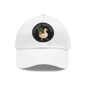 Könnte beinhalten: Eine weiße Baseballkappe mit einem runden schwarzen Aufnäher auf der Vorderseite. Der Aufnäher zeigt eine weiße Zeichentrick-Ente, die eine Holzkiste mit grünen Topfpflanzen trägt, umgeben vom Text ONE MORE WON'T HURT in weißen Buchstaben.