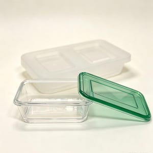 May include: A clear rectangular food storage container with a translucent green lid resting beside it, and a larger, frosted white rectangular storage container with a matching lid positioned in the background behind the smaller clear container.