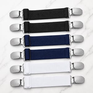 Könnte beinhalten: Ein Set aus sechs elastischen Clips mit verstellbaren Stoffbändern in Schwarz, Marineblau und Weiß. Jedes Band ist an beiden Enden mit silberfarbenen Metall-Krokodilklemmen ausgestattet, um Stofffalten zu sichern oder Kleidungsstücke zu halten.