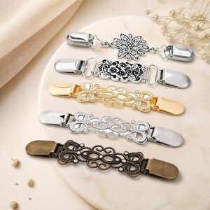 Könnte beinhalten: Fünf dekorative Cardigan-Clips mit Metallverschlüssen an beiden Enden. Die Designs umfassen eine silberne Blume, ein rechteckiges Blumenmuster, ein goldenes Filigranstück, ein silbernes Filigranstück und ein antikes bronzefarbenes Filigranstück.