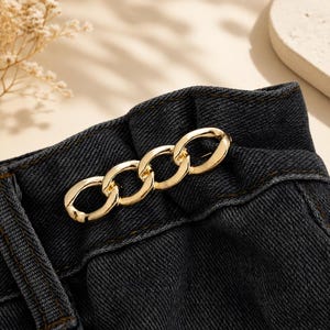 May include: A pair of dark charcoal gray denim pants featuring a high-waisted paper bag style waistband. A decorative, polished gold-tone metal chain link accent is securely attached horizontally across the front center of the gathered fabric.