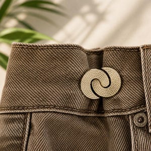 Könnte beinhalten: Eine braune Jeanshose mit hohem Bund, der mit einem dekorativen, kreisförmigen Verschluss aus goldfarbenem Metall gesichert ist. Der Verschluss besteht aus zwei ineinandergreifenden, texturierten Halbmondformen, die die Vorderseite schließen.