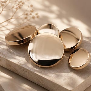 Puede incluir: Un conjunto de cinco botones circulares de metal dorado con una superficie lisa y altamente reflectante. Los botones varían ligeramente en tamaño y están dispuestos en un grupo, mostrando un acabado pulido que capta la luz con brillo.