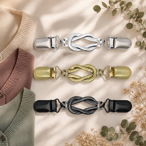 May include: Three metal cardigan clips featuring a central infinity knot design. Each clip has a polished finish in silver, gold, and matte black. The fasteners are designed with spring-loaded ends to securely grip fabric edges for garment closure.