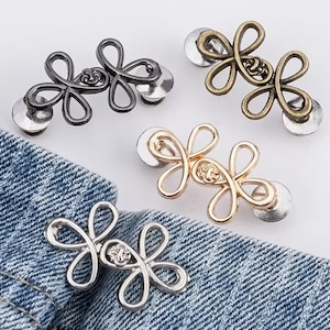 May include: Four metal fasteners with a decorative looped knot design, each featuring two circular backings. The set includes one black, one antique bronze, one gold, and one silver fastener, with the silver one attached to blue denim fabric.