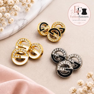 May include: Three sets of circular buttons featuring a central letter G design accented with clear rhinestones. One set is gold with black enamel, one is gold with white enamel, and one is silver with black enamel. Each button has a circular frame.