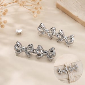 May include: Two silver-toned metal waist adjusters, each featuring a double bow design connected by a central bar. A small circular pin back is included. An inset shows the metal bow fastener attached to the waistband of light-colored denim pants.