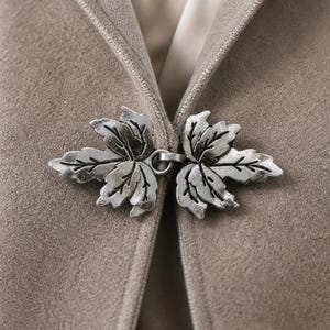 Puede incluir: Un broche de metal plateado para capa con dos diseños simétricos de hojas de arce unidos por un anillo central. El broche está sujeto a una prenda de tela beige texturizada, proporcionando un cierre seguro para los bordes delanteros.