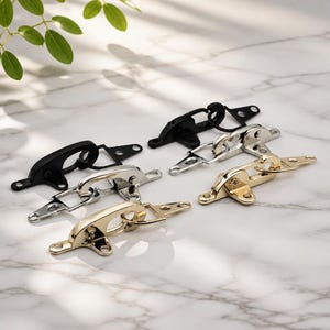 May include: A collection of six metal toggle latches arranged in pairs, featuring matte black, polished silver, and gold finishes. Each latch consists of a curved hook mechanism and a flat mounting plate with two circular screw holes for attachment.