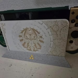 May include: A video game console dock featuring a white front panel with a gold Triforce emblem and circular patterns. The dock is paired with gold and black controllers, and a small yellow Triforce icon is centred on the grey base stand.
