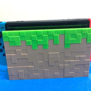 Può includere: Una base di ricarica per console di gioco con un design a blocchi pixelati, caratterizzata da uno strato superiore verde e uno inferiore marrone. Il supporto ha la forma di un blocco di terra e ospita una console nera con controller blu e rosso.