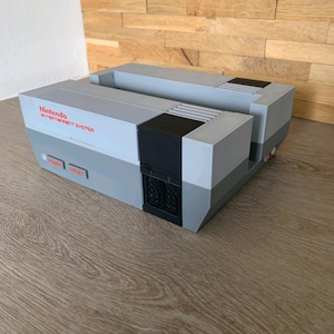May include: A grey and black Nintendo Entertainment System console model constructed from building blocks. The front features the red Nintendo Entertainment System logo, a power button, a reset button, and two controller ports labelled 1 and 2.