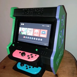 Può includere: Un cabinato arcade da tavolo nero, verde e viola che ospita una console Nintendo Switch. L'unità presenta uno schermo che mostra il menu principale, un controller Joy-Con rosa montato al centro e un controller Joy-Con blu alla base.