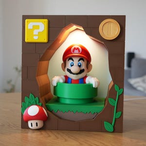 Może przedstawiać: Kwadratowa lampa z brązowym wzorem cegieł i podświetlanym wycięciem pośrodku. Wewnątrz figurka Mario wyłania się z zielonej rury. Rama zawiera żółty blok z pytajnikiem, złotą monetę, czerwonego grzyba oraz zieloną pnącą roślinę.