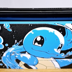 May include: A portable gaming console featuring a black screen and detachable controllers in neon blue and neon red. A rectangular dock cover is attached to the front, displaying a graphic of a blue turtle character surrounded by water bubbles.