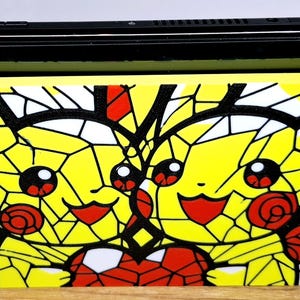 Könnte beinhalten: Eine Nintendo Switch Konsole mit einer individuellen Dockingstation-Abdeckung, die zwei gelbe Pikachu-Figuren mit einem roten Herz im Buntglas-Stil zeigt. Die Konsole hat einen hellblauen linken und einen neonroten rechten Controller.