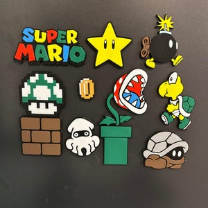 Peut inclure: Un ensemble de dix breloques en caoutchouc sur le thème de Super Mario comprenant le logo Super Mario, une étoile jaune, un Bob-omb noir, un champignon pixelisé vert et blanc sur une brique brune, une pièce d'or, un Blooper blanc, une plante Piranha, un Koopa Troopa et une carapace.