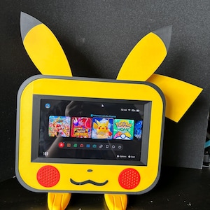 Könnte beinhalten: Eine gelb-graue Spielekonsolen-Dockingstation in Pikachu-Form mit zwei großen Ohren, einem Bildschirm mit Nintendo Switch-Menü, zwei roten runden Lautsprechern und einer schwarzen Mundgrafik auf der Vorderseite sowie zwei kleinen gelben Füßen.