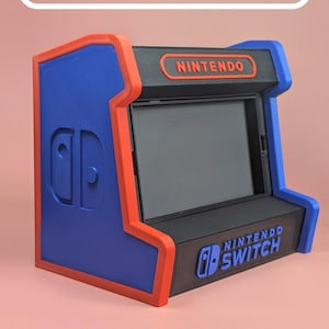 Puede incluir: Una base de carga estilo arcade azul y roja para consola Nintendo Switch. La unidad presenta un panel frontal negro con el logotipo de Nintendo en la parte superior y el logotipo de Nintendo Switch en la base. El panel lateral muestra un icono de Joy-Con.