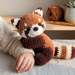 May include: A plush red panda toy made of soft, brown, white, and dark brown yarn. The stuffed animal features a round head with pointed ears, black button eyes, a dark brown nose, and a long, striped tail with alternating brown and white bands.