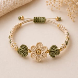 Puede incluir: Una pulsera de macramé con una flor central de color crema con una cuenta dorada en el centro, flanqueada por dos nudos en forma de hoja verde. La banda está hecha de cordón crema y verde con pequeñas cuentas doradas y un cierre corredizo.