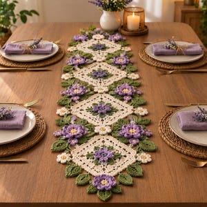 Peut inclure: Un chemin de table en crochet composé d'une série de motifs carrés couleur crème reliés par des feuilles vertes et des fleurs violettes. Le chemin est centré sur une table en bois avec quatre couverts, chacun incluant une assiette, une serviette violette et une fourchette.