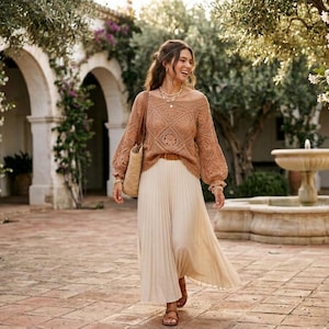 May include: A long-sleeved, tan crochet sweater with a square-patterned knit design and a wide neckline, paired with a floor-length, cream-colored pleated skirt and a woven straw shoulder bag, accessorized with a layered necklace and brown sandals.