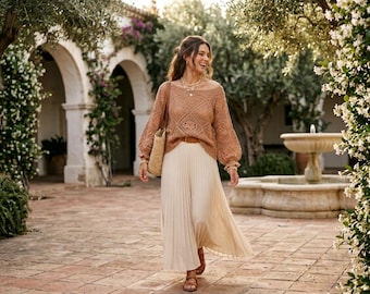 May include: A long-sleeved, tan crochet sweater with a square-patterned knit design and a wide neckline, paired with a floor-length, cream-colored pleated skirt and a woven straw shoulder bag, accessorized with a layered necklace and brown sandals.