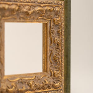 Puede incluir: Un marco de fotos cuadrado que presenta un patrón de relieve dorado ornamentado con intrincados motivos florales y de volutas. El borde exterior del marco muestra un acabado verde oscuro texturizado, que contrasta con el interior de oro metálico.