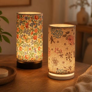 May include: Two cylindrical table lamps with illuminated fabric shades. The left lamp features a dense, colorful floral pattern with red, yellow, and blue blossoms. The right lamp has a beige shade with a sparse, delicate botanical print.