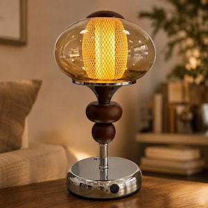 Puede incluir: Una lámpara de mesa con una pantalla de cristal redonda en tono ámbar y un cilindro central de malla dorada. La base consta de dos esferas de madera marrón oscuro sobre un soporte de cromo pulido y una base circular con un botón de encendido.