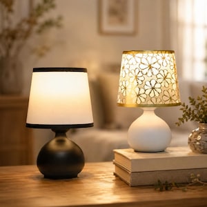 May include: Two table lamps with rounded bases. The left lamp has a matte black base and a plain white tapered shade with black trim. The right lamp has a white base and a gold-toned tapered shade featuring a repeating white floral pattern.