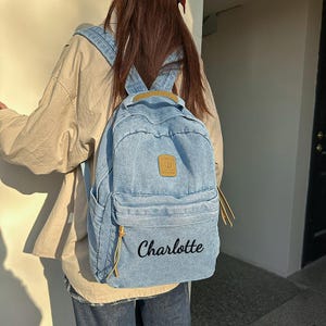 Puede incluir: Una mochila de mezclilla azul claro con un parche rectangular color canela en el bolsillo superior y el nombre Charlotte bordado en letra cursiva negra en el bolsillo frontal inferior. Incluye dos bolsillos laterales y tiradores de cremallera.