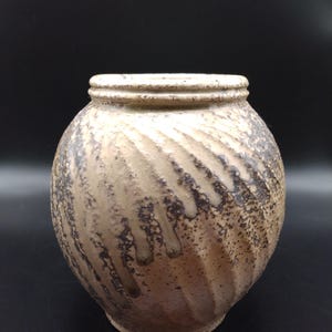 Könnte beinhalten: Eine runde Keramikvase mit einer strukturierten, beigen Oberfläche und diagonalen, spiralförmigen Rillen. Die Außenseite zeigt eine gesprenkelte, dunkelbraune und graue Glasur, die an den Seiten herabläuft und in einem erhöhten Rand endet.
