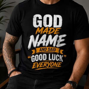 Puede incluir: Una camiseta negra de manga corta con texto blanco y naranja de estilo desgastado que dice: GOD MADE NAME AND SAID GOOD LUCK EVERYONE. El diseño utiliza una mezcla de letras mayúsculas en negrita y fuentes cursivas con un elemento gráfico de pancarta.