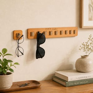 Peut inclure: Un organisateur mural rectangulaire en bois avec dix boucles en cuir pour suspendre des lunettes, portant le texte gravé "Sophia" et une icône de lunettes. Un crochet carré en bois séparé supporte une paire de lunettes fines.