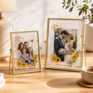 May include: Two gold-toned metal floating picture frames with glass panes. The frames feature white line-art floral borders and pressed sunflower accents. One frame displays a family portrait, while the other shows a wedding couple.