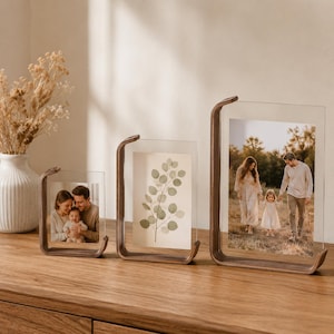 Può includere: Tre cornici portafoto fluttuanti con basi curve in legno scuro e pannelli in acrilico trasparente. Le cornici contengono fotografie di una famiglia, un'illustrazione botanica di foglie di eucalipto e una famiglia che cammina in un campo.