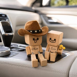 Könnte beinhalten: Zwei kleine Holzfiguren in Würfelform mit beweglichen Gliedmaßen. Eine Figur trägt einen braunen Filz-Cowboyhut mit einem schwarzen, sternbesetzten Band und ist mit dem Namen Ethan graviert. Die andere trägt den Namen Luna und hält gelbe Blumen.