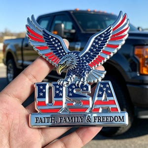 Könnte beinhalten: Ein metallisches Emblem mit einem Weißkopfseeadler mit ausgebreiteten Flügeln in den Farben Rot, Weiß und Blau. Unter dem Adler sind die Buchstaben USA in einem Flaggenmuster dargestellt. Ein horizontaler Balken trägt die Aufschrift FAITH, FAMILY & FREEDOM.