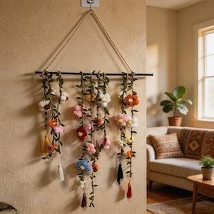 May include: A wall hanging featuring a black horizontal rod suspended by a jute rope. Three vertical strands of green faux vines are adorned with multi-colored crocheted flowers in shades of white, pink, red, orange, yellow, and blue, with tassels.