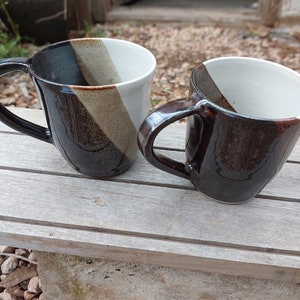 Può includere: Due tazze in ceramica con smalto nero, bianco e marrone. Le tazze hanno un manico e sono appoggiate su una superficie di legno.