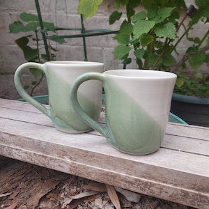 Peut inclure: Deux tasses en céramique avec un glaçage vert et blanc. Les tasses ont une forme arrondie et une poignée de chaque côté.