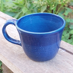 Könnte beinhalten: Eine tiefblaue Keramiktasse mit abgerundeter Form und einem Henkel. Die Tasse hat eine gesprenkelte Glasur und steht auf einer Holzoberfläche.