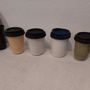 May include: A collection of ceramic travel mugs with black and blue silicone lids. The mugs vary in color, including beige, white, and green. The mugs are designed to hold hot beverages and are suitable for travel.